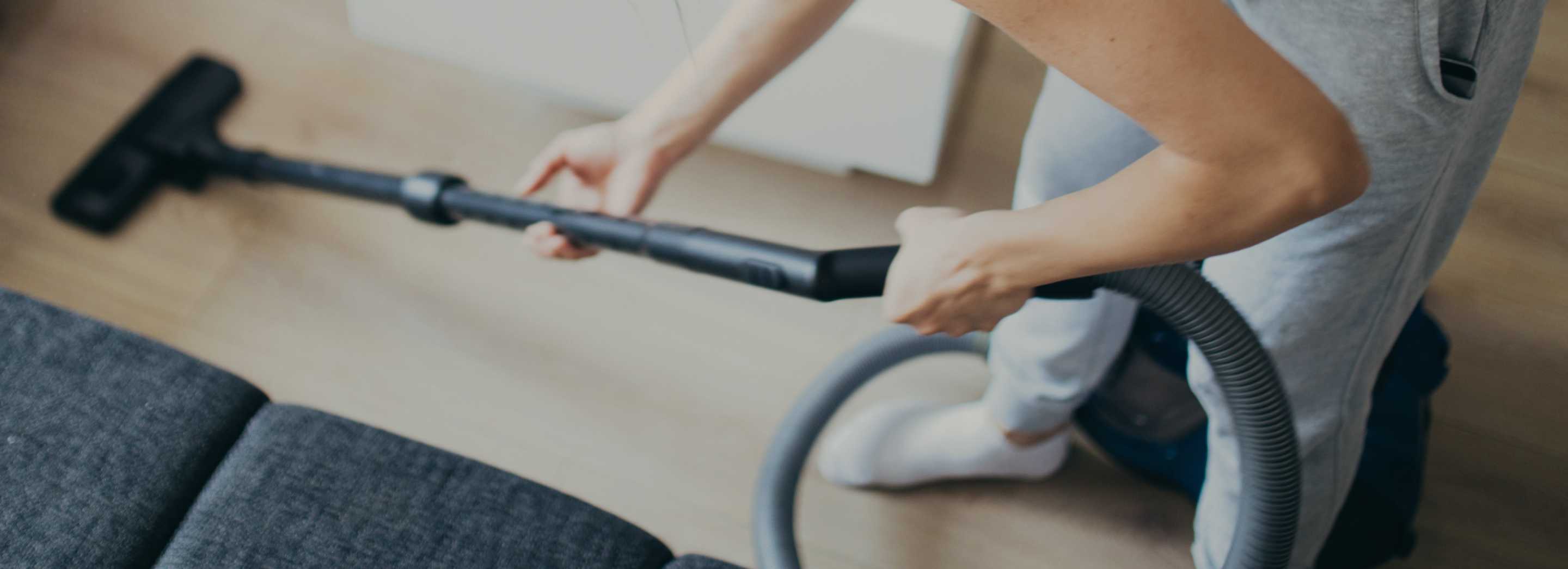 Someone with a long arm vacuum and head is shown vacuuming their wooden floors in a pair of grey pants
