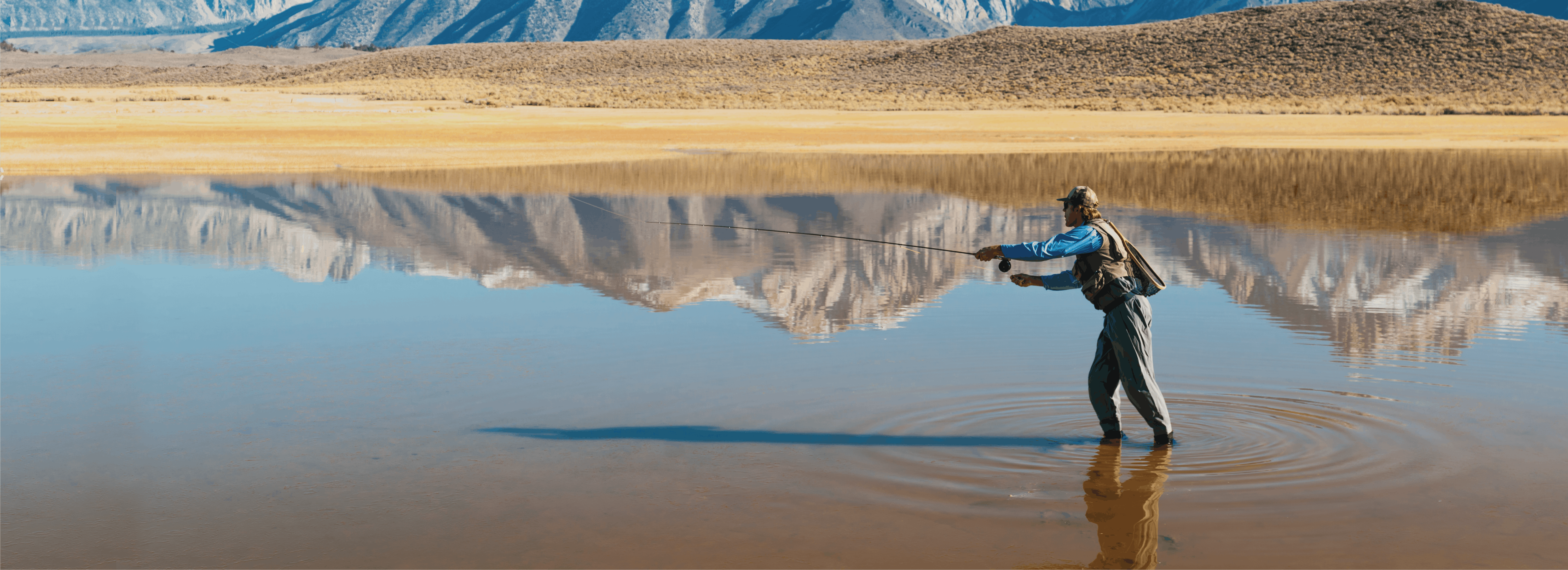 A man casts his reel in a lake reflecting snowy mountains. 